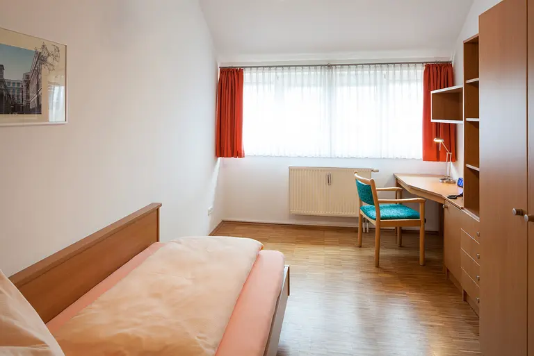 Schlafzimmer mit Bett, Schreibtisch, Schränken und einem großen Fenster