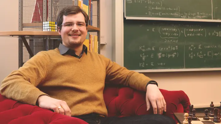 Tobias Ried sitzt in einem Sessel, daneben eine Tafel mit mathematischen Formeln