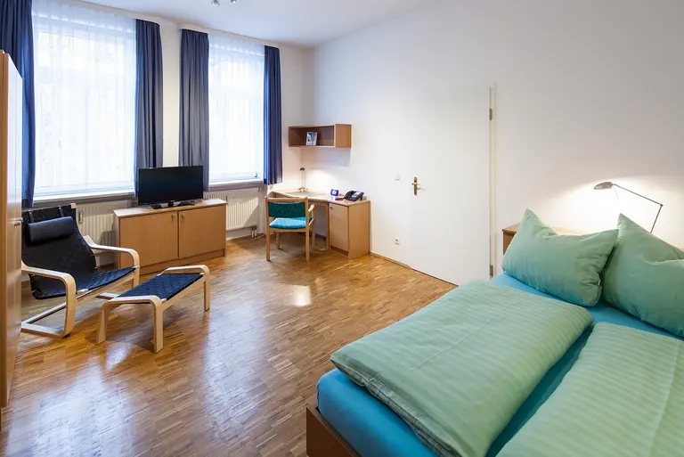 Zimmer mit Doppelbett, Sessel, Schrank, Schreibtisch und Fernseher