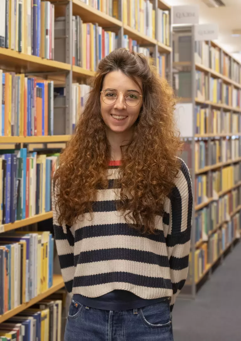 Portrait of Lisa Seccia with arms behind her back in front of bookshelves